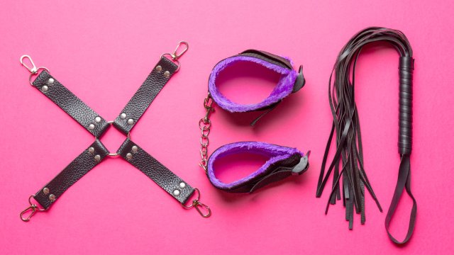 Akcesoria BDSM – kajdanki, pejcz i knebel na czerwonym tle